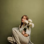 A serene woman sitting on a chair holding white flowers in a studio setting.